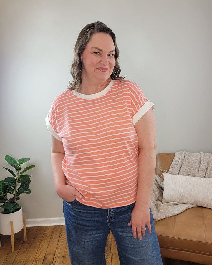 Wearing the ZENANA Brealyn Stripe Tee, a woman in blue jeans stands indoors, smiling with one hand in her pocket. A plant and tan couch with a blanket and pillow are in the background. This tee combines lightweight comfort and style.