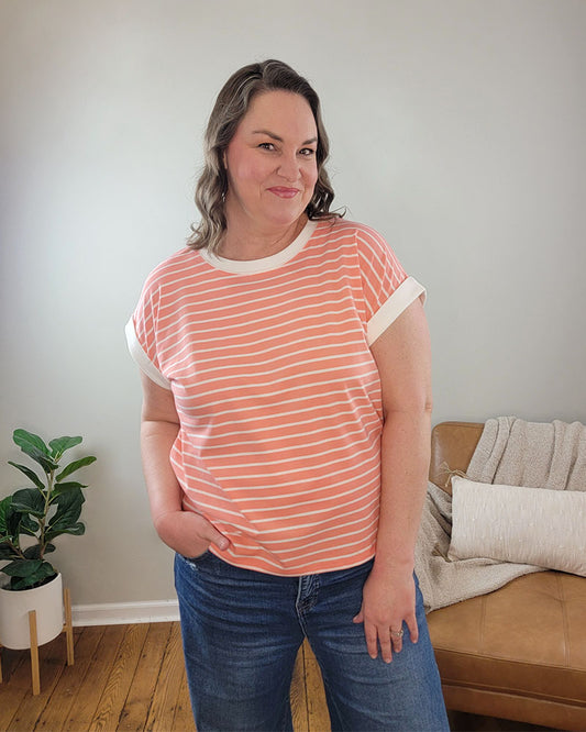 Wearing the ZENANA Brealyn Stripe Tee, a woman in blue jeans stands indoors, smiling with one hand in her pocket. A plant and tan couch with a blanket and pillow are in the background. This tee combines lightweight comfort and style.