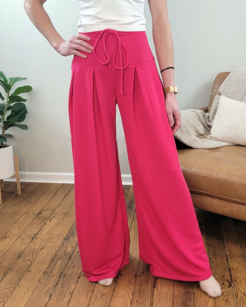 Indoors on a wooden floor, someone wears Selfmade Orange Melanie Pleated Pants—high waist, wide leg, and tie waistband—ideal for travel. A plant, tan couch, and beige blanket complete the background.