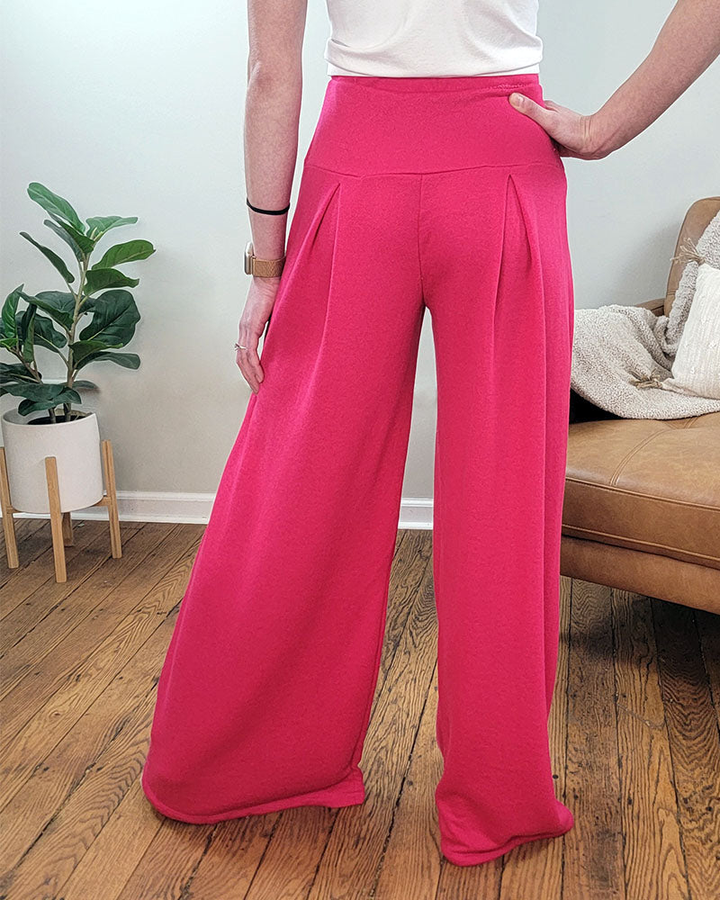 Indoors on a wooden floor, a person models Selfmade Orange's Melanie Pleated Pants in bright pink with a white top. One hand rests on their hip. A green potted plant and tan couch with blanket complete the scene.