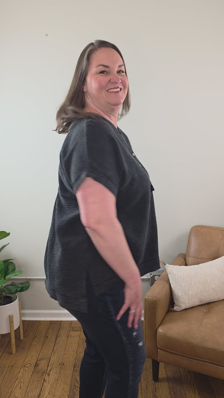 A woman with straight, shoulder-length hair wears the ZENANA Kasey Short Sleeve Washed Henley Shirt and jeans, available at Cosmos and Clover Boutique, smiling in a living room featuring a plant, brown leather couch, and white pillow in the background.