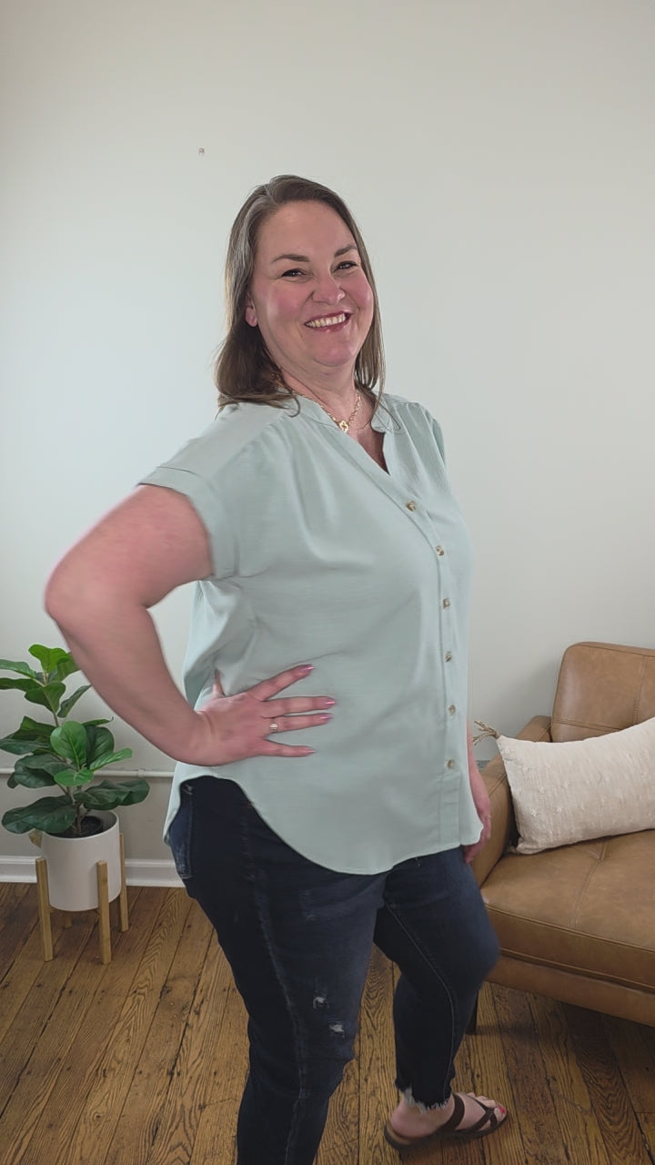 A woman with shoulder-length brown hair stands indoors in the Kayla Rolled Sleeve Button Down Top from Cosmos and Clover Boutique, light blue, paired with dark jeans. A brown sofa, white pillow, and potted plant emphasize this versatile blouse for any casual setting.