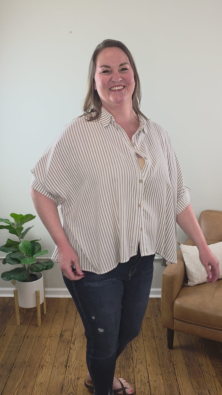 A woman with long brown hair smiles indoors beside a tan armchair and potted plant, wearing the Ryan Striped Short Sleeve Button Down Shirt and dark jeans, both from Cosmos and Clover Boutique in a light-colored room with wooden floors.