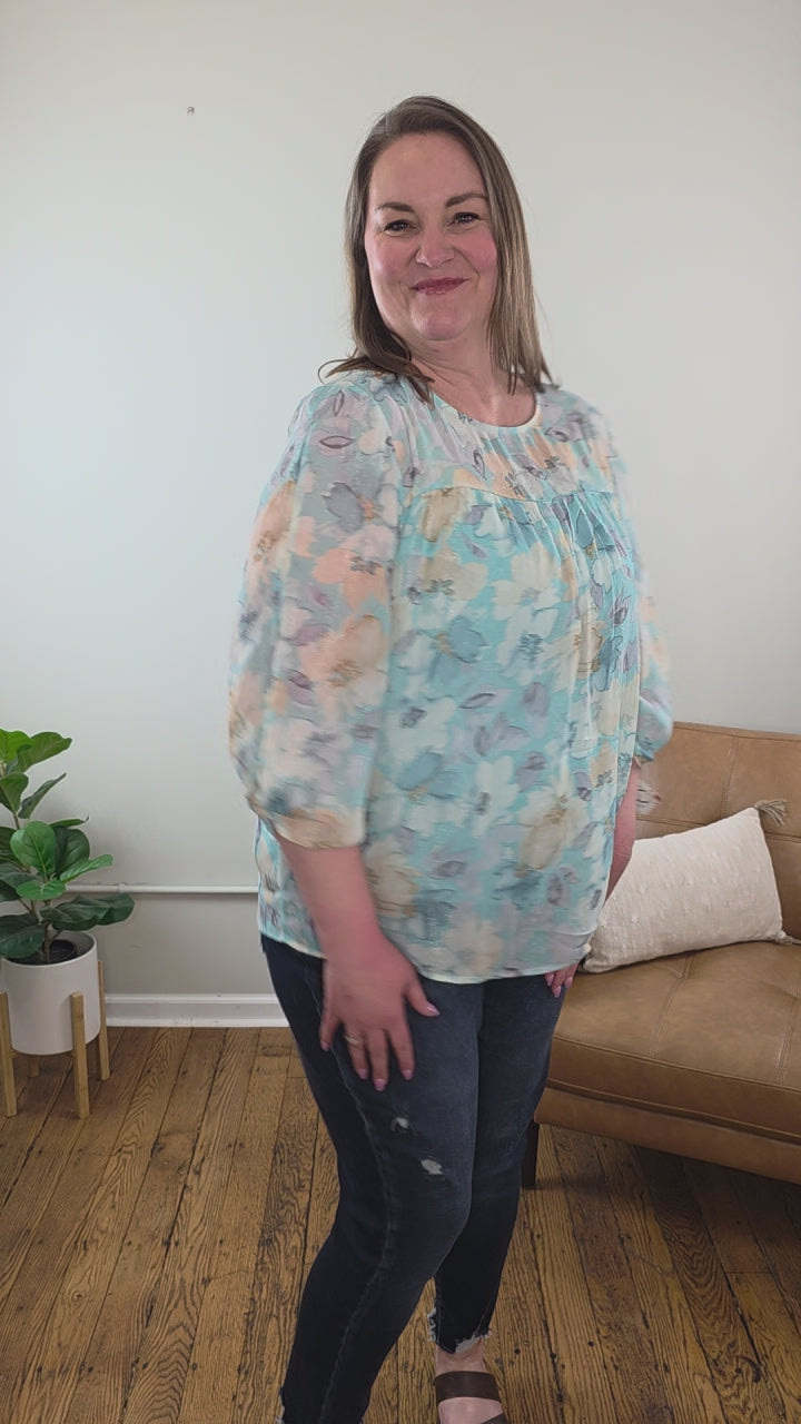 A woman with long hair stands smiling in a room, wearing the Kaley Floral 3/4 Sleeve Blouse with Sheer Overlay and dark jeans, both from Cosmos and Clover Boutique. The wooden-floored space features a tan couch, potted plant, and a plain light wall.