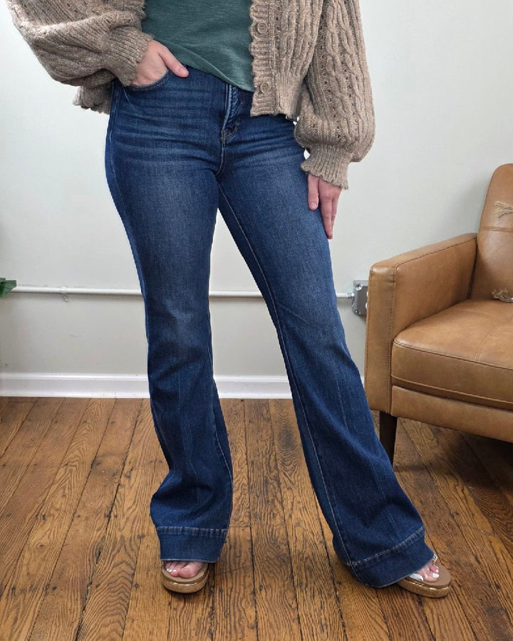 A person stands on a hardwood floor next to a tan chair, their left hand resting on their hip, wearing a chunky beige sweater, green shirt, and the Rachel High Rise Trouser Flares from Risen with Tummy Control in a classic blue wash.