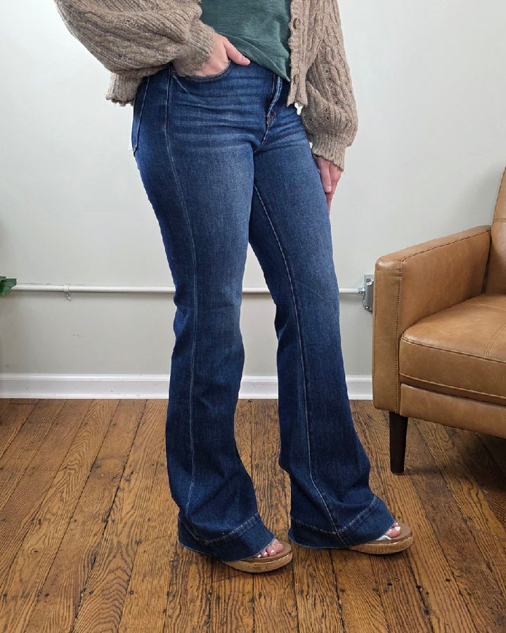 A person stands on a wooden floor wearing the Rachel High Rise Trouser Flares with Tummy Control from Risen in a blue wash, styled with a green top, beige knit sweater, and brown platform sandals beside a tan armchair against a light-colored wall.