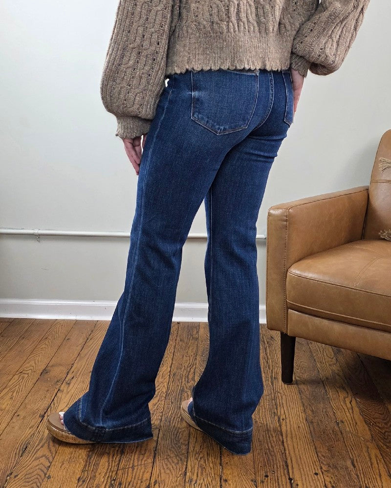 Wearing the Rachel High Rise Trouser Flares from Risen with Tummy Control in a dark wash, a beige chunky knit sweater with balloon sleeves, and brown sandals, a person stands on a wooden floor next to a brown armchair.