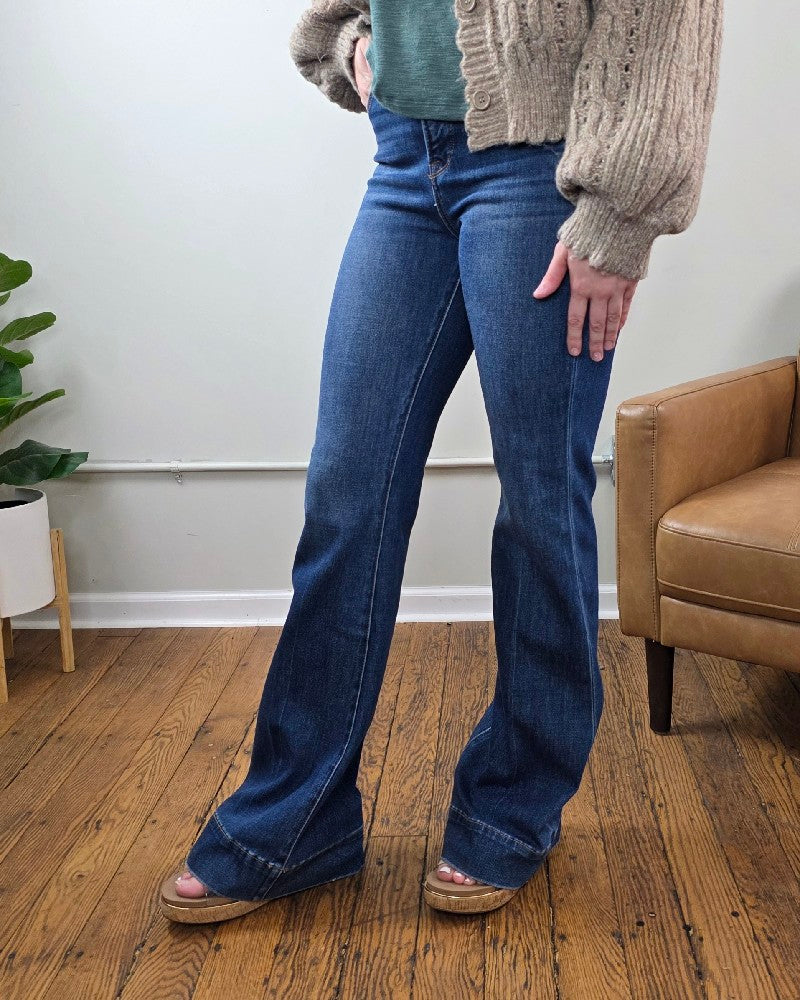Wearing the Rachel High Rise Trouser Flares from Risen with Tummy Control, paired with a green top and a beige knit cardigan, a person stands on a wooden floor next to a tan chair and a potted plant.