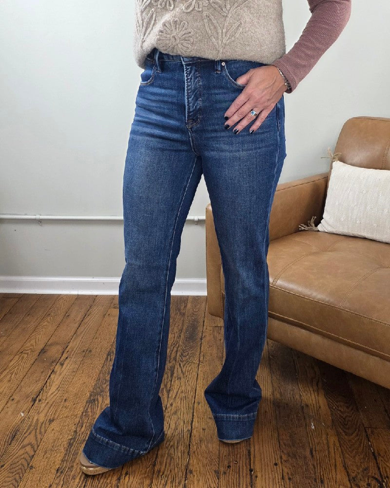 A person stands indoors on a wooden floor, wearing the Rachel High Rise Trouser Flares from Risen with Tummy Control in classic blue denim, paired with a beige sweater and a mauve long-sleeve shirt. One hand rests on their hip near a tan couch topped with a white pillow.