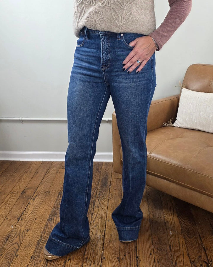A person stands indoors on a wooden floor, wearing the Rachel High Rise Trouser Flares from Risen with Tummy Control in classic blue denim, paired with a beige sweater and a mauve long-sleeve shirt. One hand rests on their hip near a tan couch topped with a white pillow.