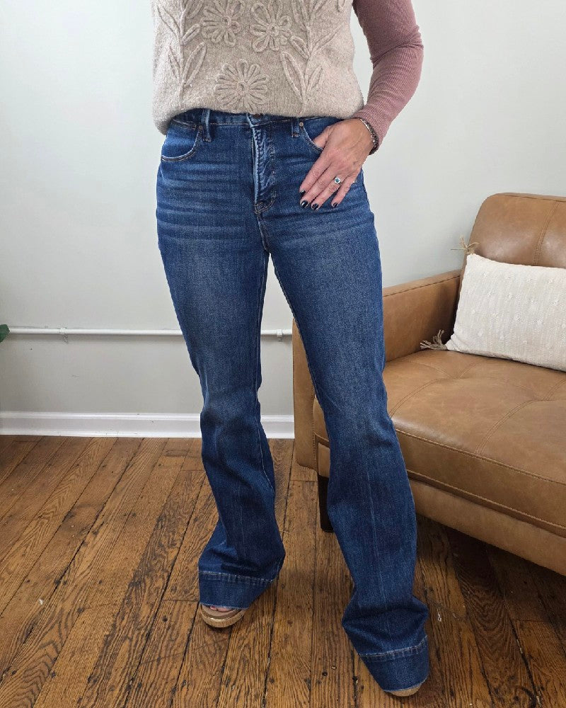 A person is standing indoors wearing the Rachel High Rise Trouser Flares from Risen with Tummy Control in a blue wash, styled with a beige sleeveless floral top layered over a long-sleeve pink shirt. A wooden floor and a brown couch with a pillow are visible in the background.