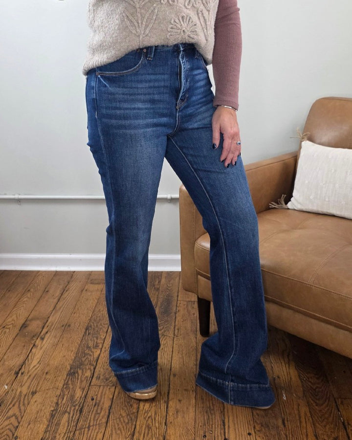 Wearing the Rachel High Rise Trouser Flares from Risen with Tummy Control and a beige sweater, a person stands next to a brown leather chair on a wooden floor, with their face not visible.