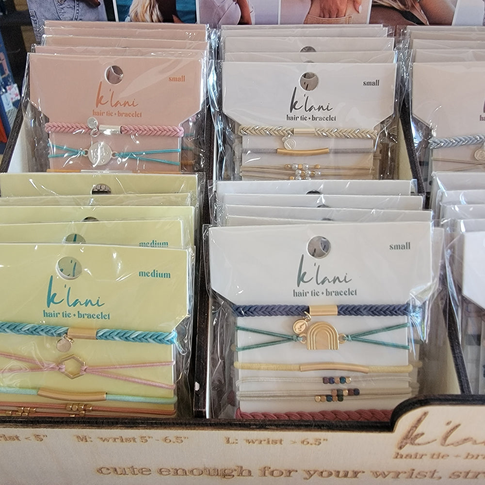 K'lani Wholesale displays K'lani Hair Tie Bracelet Sets in pastel hues, sorted by small and medium sizes. Each set features several hair ties with charming accents including rainbows and geometric designs.