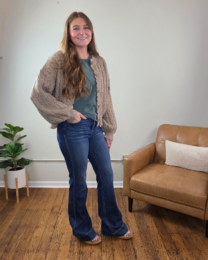 A woman with long brown hair stands indoors on wooden floors, wearing the blu pepper Rosa Eyelet Cable Knit Scalloped Trim Cardigan, a green top, blue flared jeans, and sandals. A small plant and tan armchair are behind her.