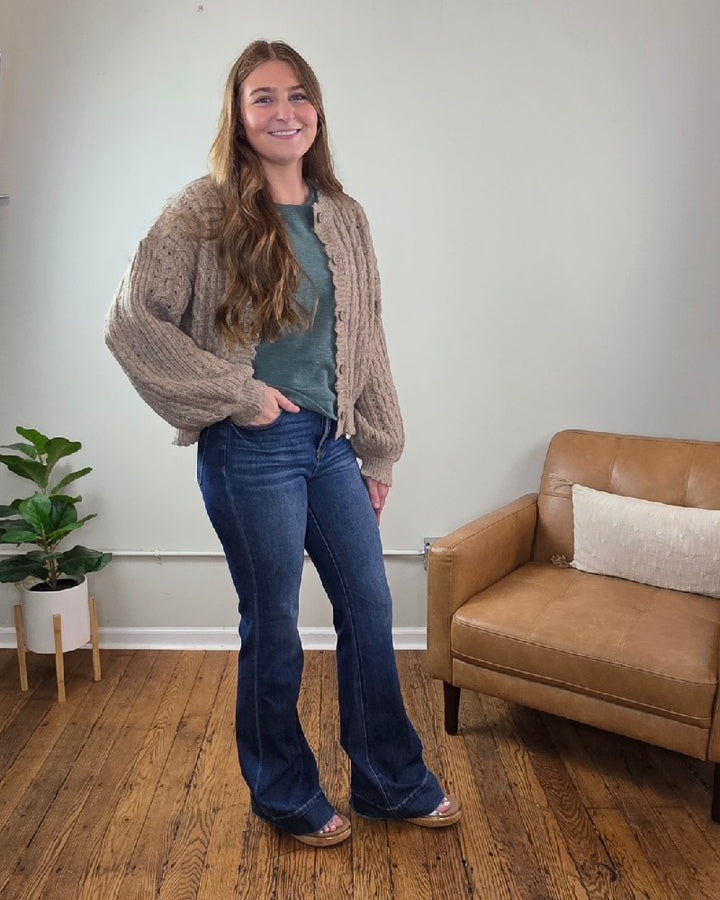 A woman with long brown hair stands indoors on wooden floors, wearing the blu pepper Rosa Eyelet Cable Knit Scalloped Trim Cardigan, a green top, blue flared jeans, and sandals. A small plant and tan armchair are behind her.
