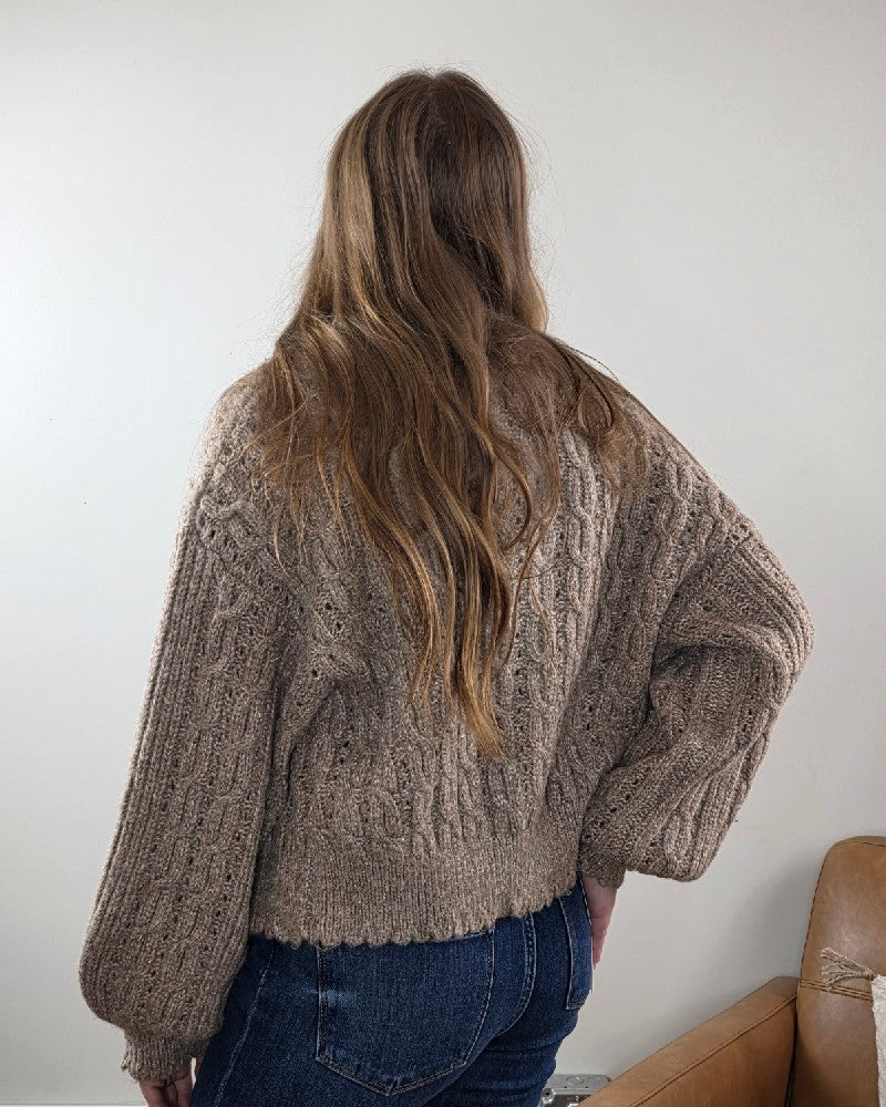 A person with long, wavy brown hair wears the blu pepper Rosa Eyelet Cable Knit Scalloped Trim Cardigan and blue jeans, standing with their back to the camera in a neutral-colored room.