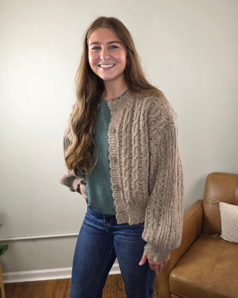 A young woman with long brown hair, in a green top, jeans, and the blu pepper Rosa Eyelet Cable Knit Scalloped Trim Cardigan, stands smiling in a living room beside a light brown couch.