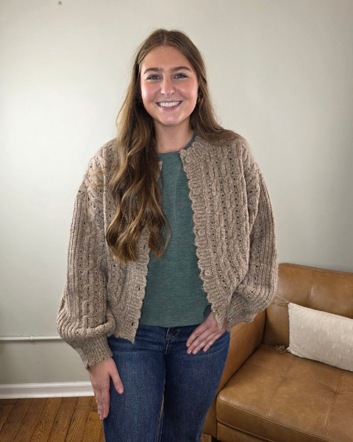 A woman with long brown hair smiles in a room, wearing the blu pepper Rosa Eyelet Cable Knit Scalloped Trim Cardigan over a green top and jeans.