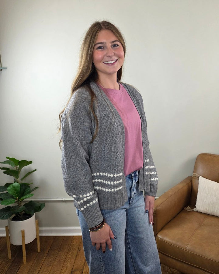 A person wearing a grey Sierra Honeycomb Textured Cardigan with white dashed-line accents on the sleeves over a pink t-shirt and blue jeans.