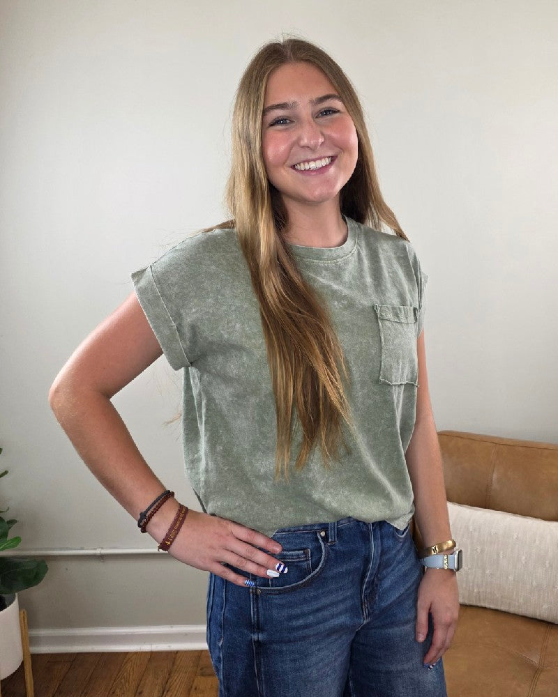A young woman with long brown hair, wearing the ZENANA Emma Snow Washed Short Sleeve Top in green—a relaxed fit t-shirt made from 100% cotton—paired with blue jeans, stands indoors smiling with one hand on her hip. In the background, there is a tan couch and a plant.