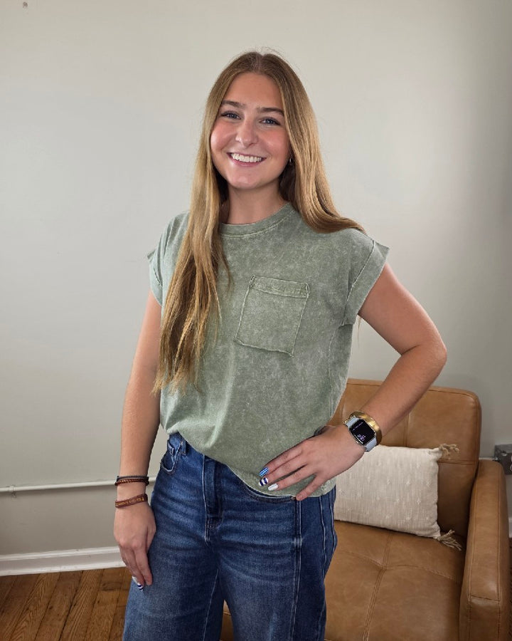 A smiling woman with long hair, wearing the ZENANA Emma Snow Washed Short Sleeve Top—a relaxed fit, 100% cotton green pocket t-shirt—and blue jeans, stands next to a tan sofa with a beige pillow in a simple, well-lit room.