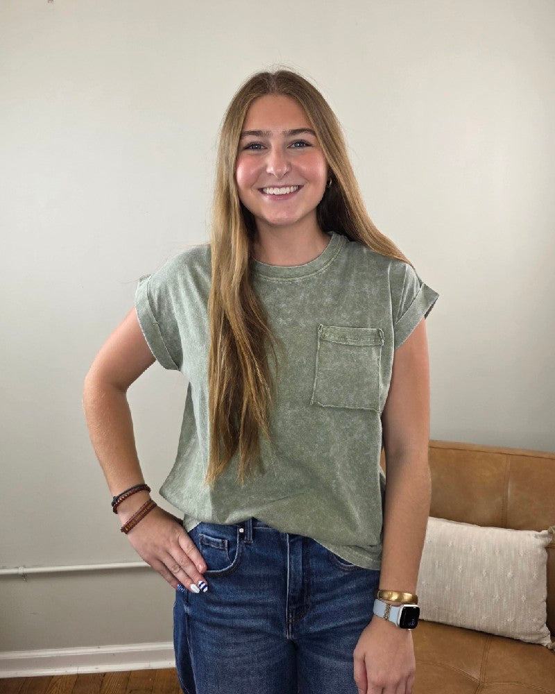 A smiling woman with long light brown hair wears the Emma Snow Washed Short Sleeve Top by ZENANA—a relaxed fit, 100% cotton green tee—paired with jeans as she stands indoors in front of a beige couch and light-colored wall.