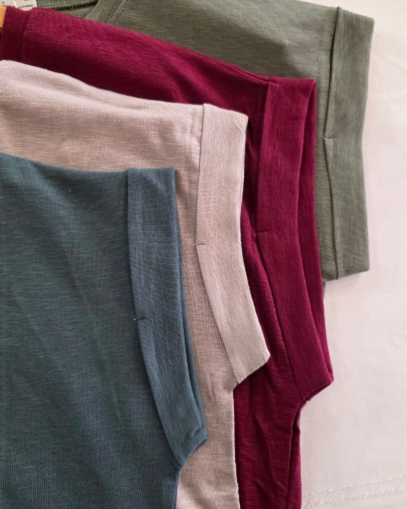 Four ZENANA Thea Rolled Sleeve Tees in green, dark pink, light beige, and blue are neatly arranged side by side on a light surface, showcasing their soft cotton-blend fabric and stylish rolled sleeves.