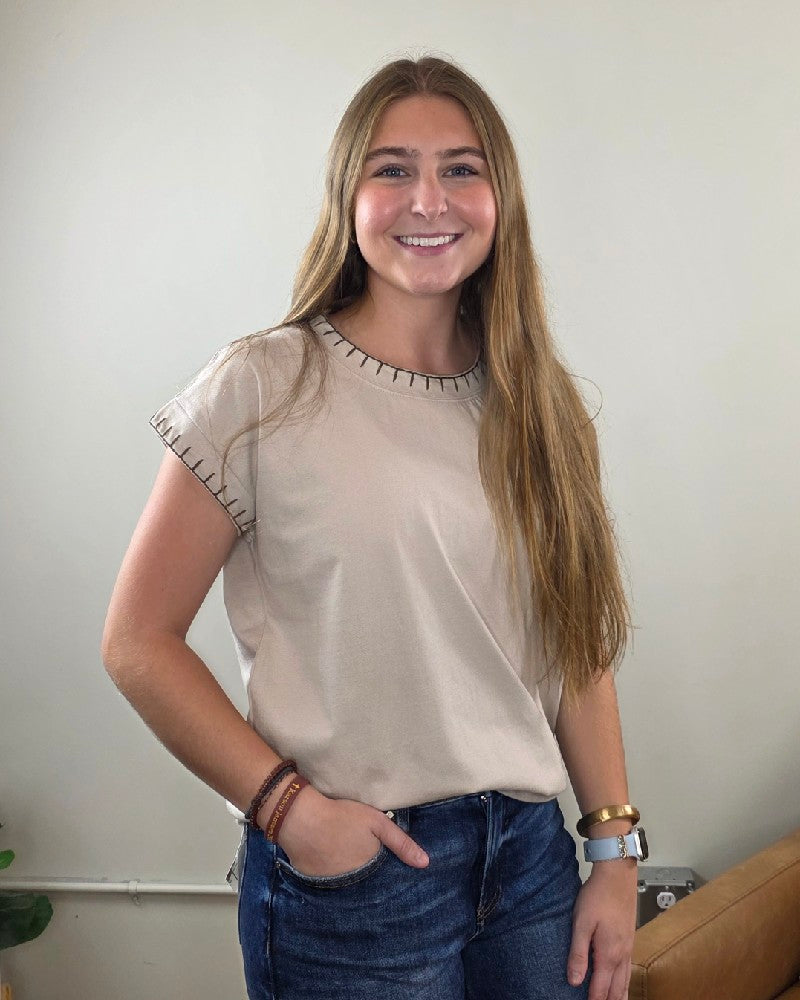 A woman with long brown hair, dressed in the ZENANA Theresa Top with Stitching Detail in sand beige and blue jeans, stands smiling with one hand in her pocket against a plain light-colored wall.