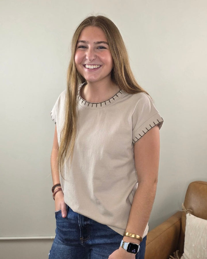 A young woman with long brown hair smiles at the camera, wearing the ZENANA Theresa Top with Stitching Detail in beige, paired with blue jeans and a smart watch. She stands in a room featuring a light wall and a brown couch.