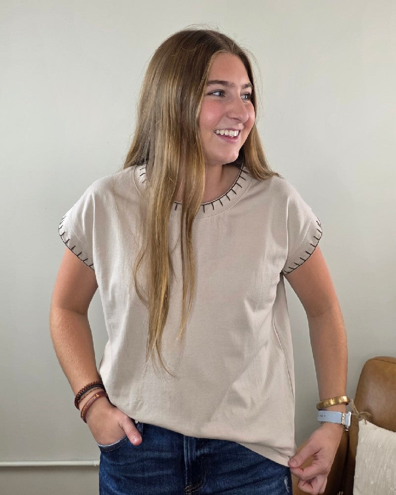 A woman with long brown hair stands smiling and looking to her right against a light background, wearing the ZENANA Theresa Top with Stitching Detail in sand beige, paired with blue jeans and bracelets.