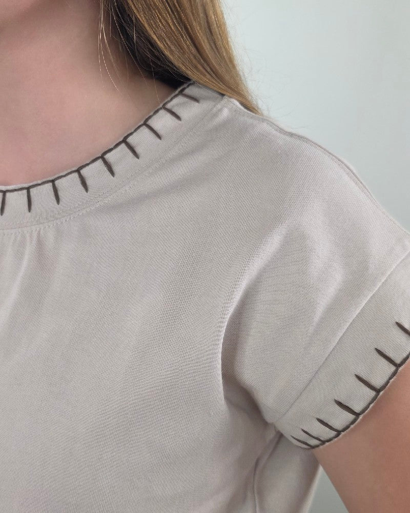 Close-up of a person wearing the ZENANA Theresa Top with Stitching Detail in sand beige, featuring dark artisan-inspired stitching along the collar and sleeve edges. Only the shoulder, neck, and part of the hair are shown, emphasizing the stylish decorative details on this short-sleeve shirt.