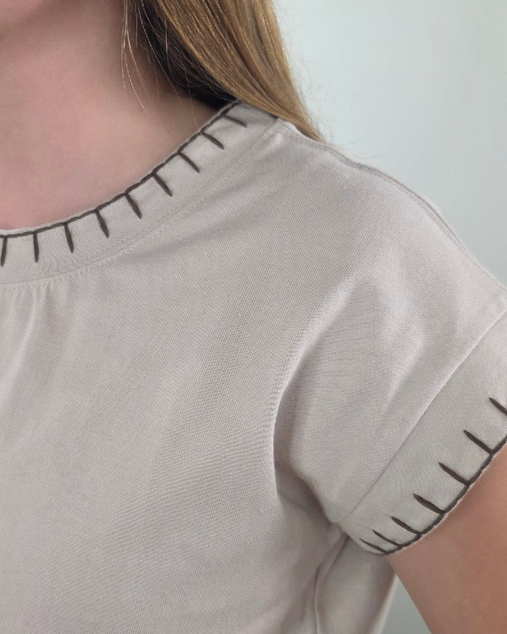 Close-up of a person wearing the ZENANA Theresa Top with Stitching Detail in sand beige, featuring dark artisan-inspired stitching along the collar and sleeve edges. Only the shoulder, neck, and part of the hair are shown, emphasizing the stylish decorative details on this short-sleeve shirt.