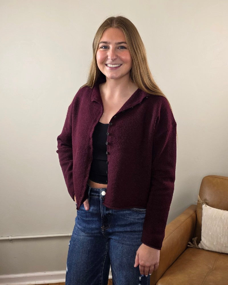 A young woman with long brown hair smiles indoors, wearing the blu pepper Violet Oxblood Button Down Cardigan over a black crop top and blue jeans, with a tan couch and cream wall in the background.