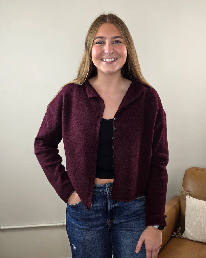 A young woman with long light brown hair smiles indoors, wearing the blu pepper Violet Oxblood Button Down Cardigan over a black top and blue jeans, with one hand in her pocket.