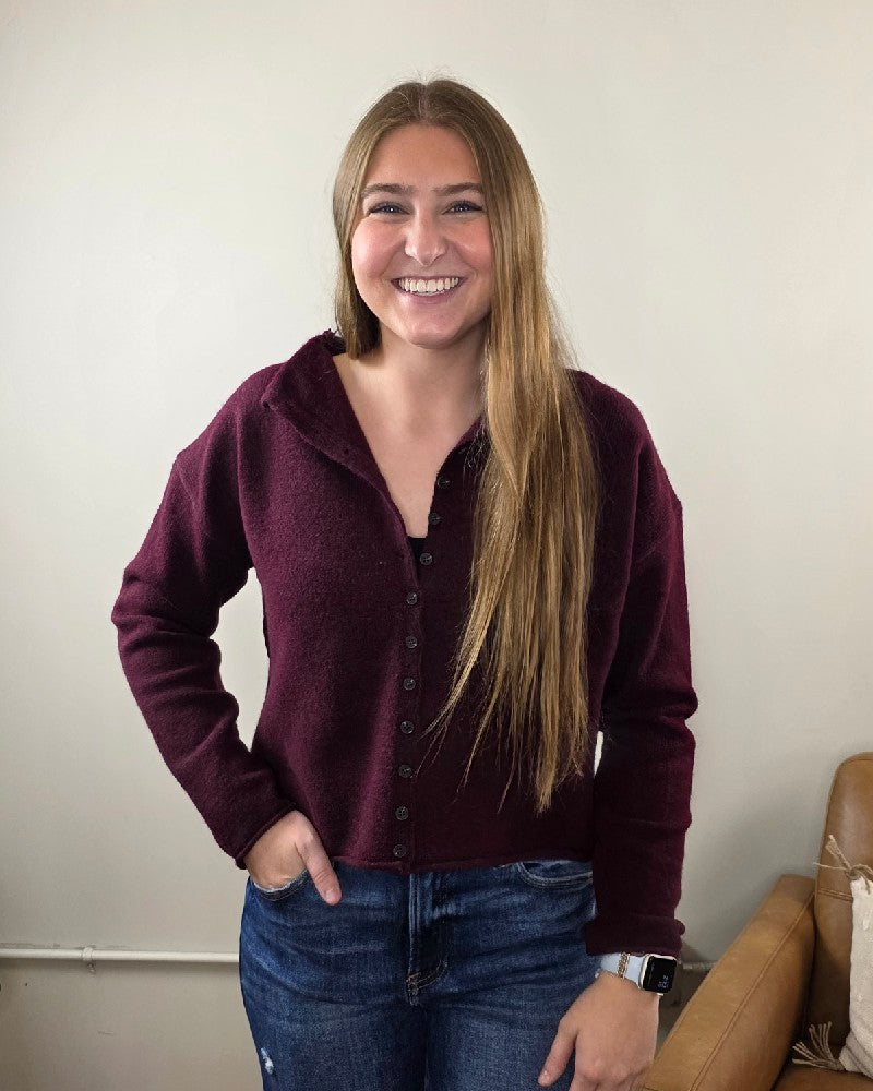 A person with long blonde hair smiles at the camera indoors, wearing the blu pepper Violet Oxblood Button Down Cardigan and blue jeans, with one hand in their pocket. A brown couch appears partially on the right.