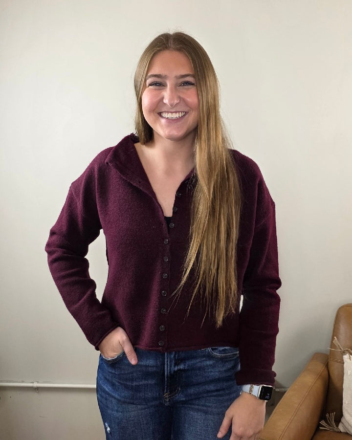 A person with long blonde hair smiles at the camera indoors, wearing the blu pepper Violet Oxblood Button Down Cardigan and blue jeans, with one hand in their pocket. A brown couch appears partially on the right.