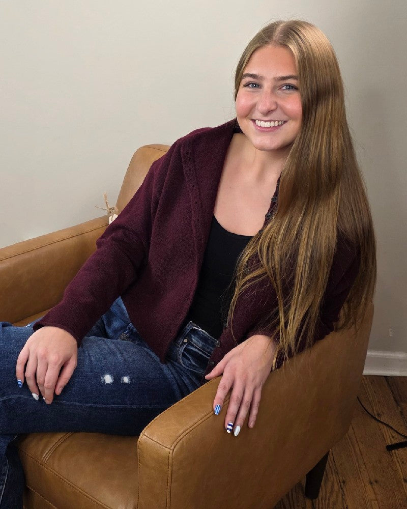 A young woman with long straight hair smiles while seated on a tan armchair, wearing the blu pepper Violet Oxblood Button Down Cardigan over a black top and ripped blue jeans. Her nails have a striped design.