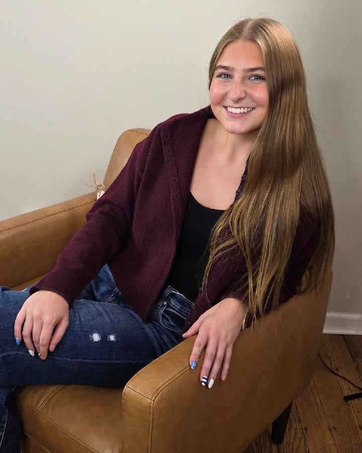 A young woman with long straight hair smiles while seated on a tan armchair, wearing the blu pepper Violet Oxblood Button Down Cardigan over a black top and ripped blue jeans. Her nails have a striped design.