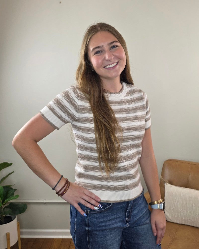 A young woman with long brown hair smiles and poses with one hand on her hip, wearing the Thread & Supply Welles Striped Short Sleeve Sweater paired with jeans in a cozy indoor space featuring neutral decor and a potted plant.