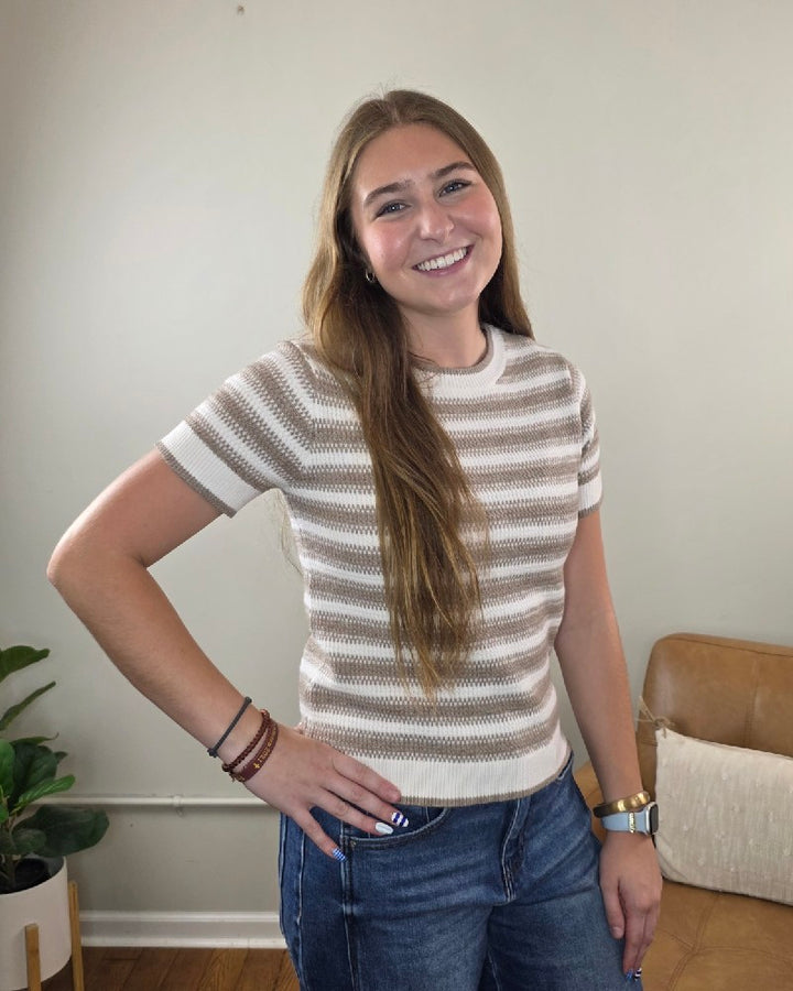 A young woman with long brown hair smiles and poses with one hand on her hip, wearing the Thread &amp; Supply Welles Striped Short Sleeve Sweater paired with jeans in a cozy indoor space featuring neutral decor and a potted plant.