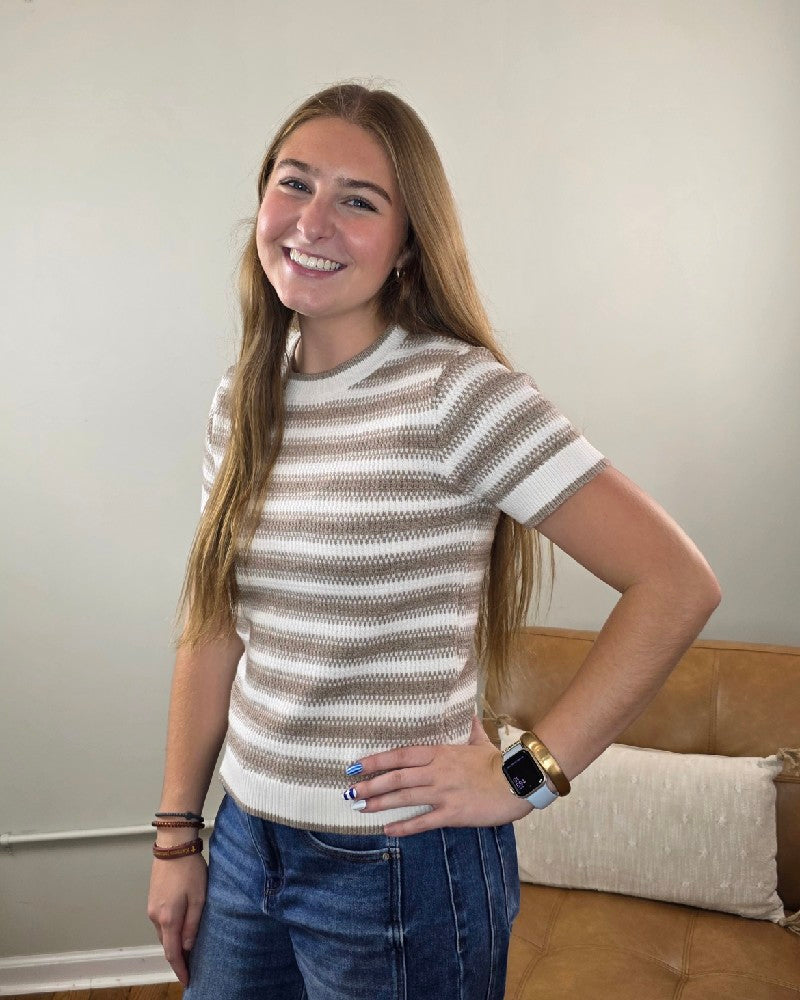 A young woman with long hair smiles, wearing the Thread & Supply Welles Striped Short Sleeve Sweater paired with jeans. Standing indoors by a brown couch with her hand on her hip, she shows off a smartwatch and bracelets—a stylish way to highlight your favorite transitional fall sweater.