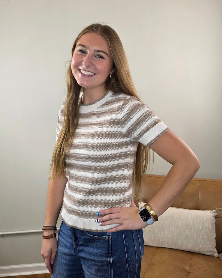 A young woman with long hair smiles, wearing the Thread &amp; Supply Welles Striped Short Sleeve Sweater paired with jeans. Standing indoors by a brown couch with her hand on her hip, she shows off a smartwatch and bracelets—a stylish way to highlight your favorite transitional fall sweater.