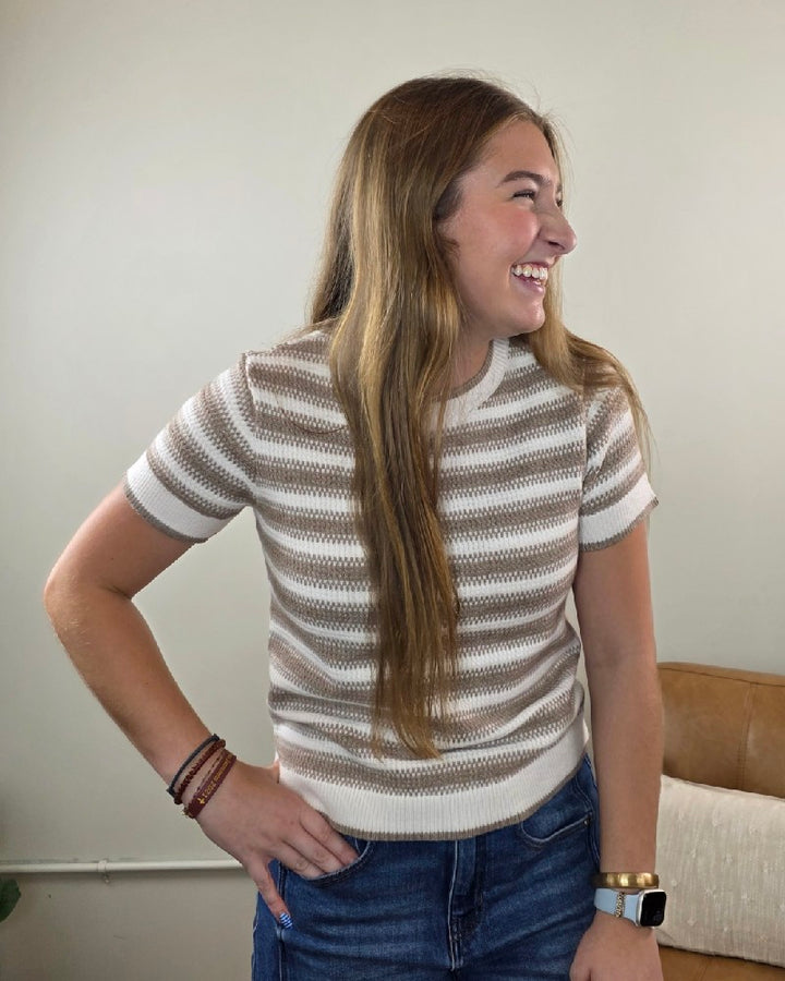 A woman with long brown hair smiles while looking to the side. She wears the Thread &amp; Supply Welles Striped Short Sleeve Sweater, paired with blue jeans, bracelets, and a smartwatch, standing indoors against a neutral background.