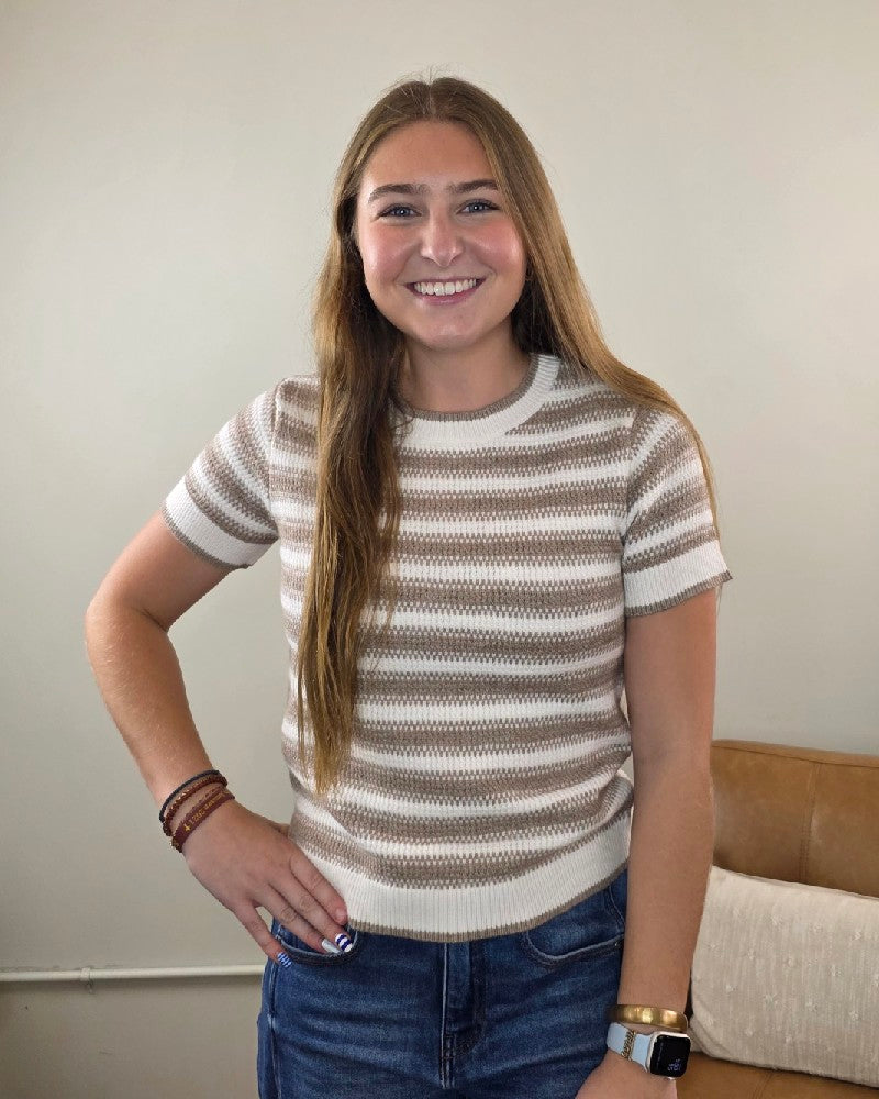 A young woman with long brown hair stands smiling, wearing the Thread &amp; Supply Welles Striped Short Sleeve Sweater paired with blue jeans, bracelets, and a watch. She poses indoors with one hand on her hip in front of a beige wall and couch.