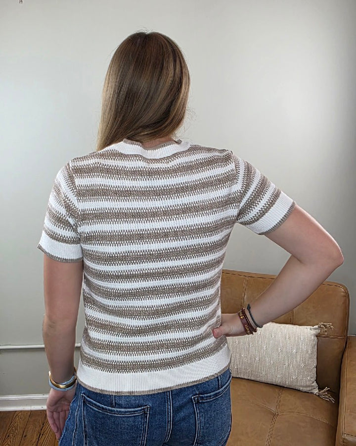 A person with long hair stands with their back to the camera, hand on hip, showcasing the Welles Striped Short Sleeve Sweater in beige and white by Thread &amp; Supply. This transitional fall piece combines comfort and style, set against a neutral backdrop with a tan leather chair.