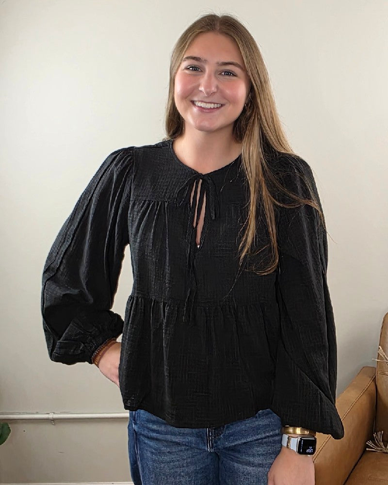 A young woman with long brown hair, smiling and standing indoors in front of a beige wall, is wearing blue jeans and a stylish black HYFVE, Inc Willa Tie Front Peplum Blouse with long sleeves.