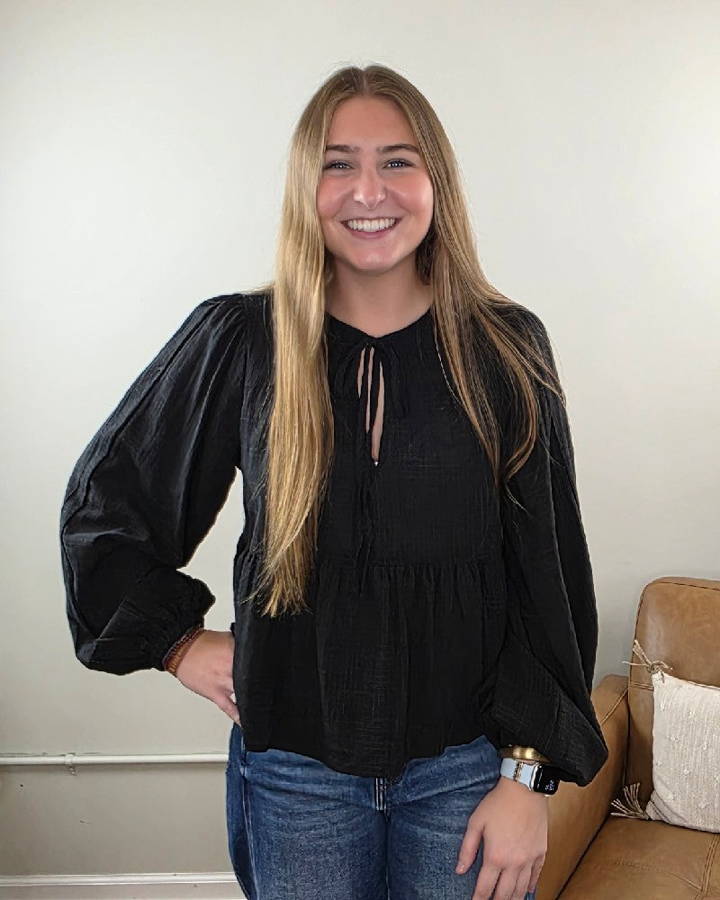 A young woman with long, straight hair stands indoors beside a brown couch and a light-colored wall, smiling at the camera while wearing blue jeans and a black Willa Tie Front Peplum Blouse by HYFVE, Inc.