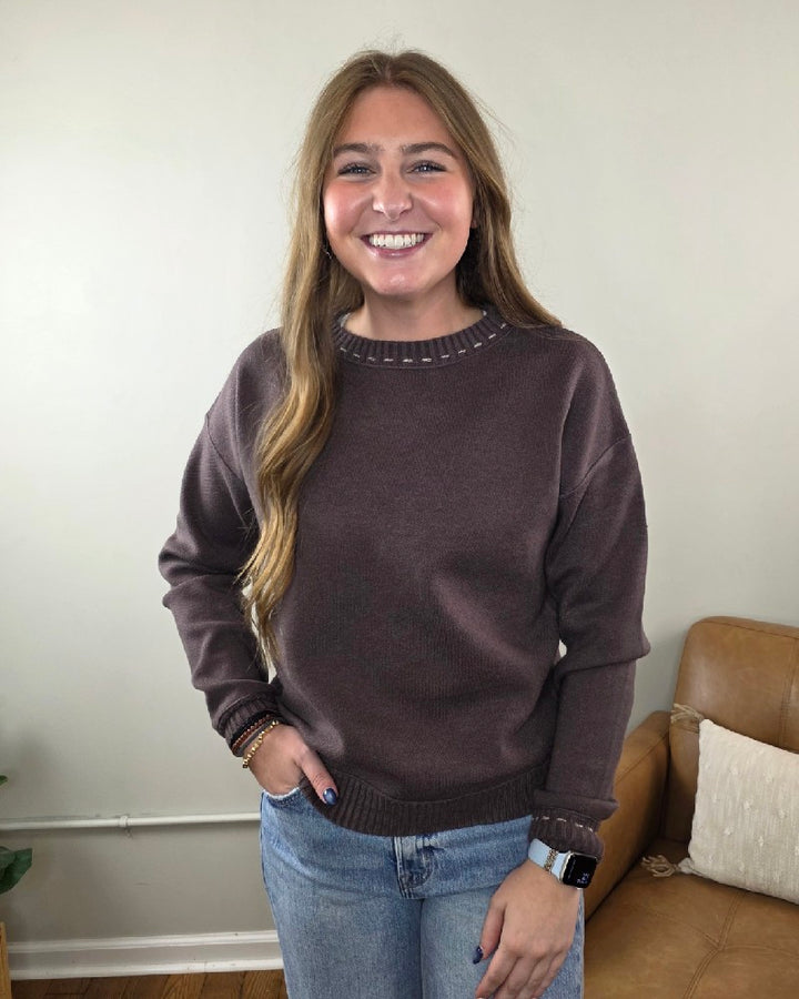 A smiling woman wears a dark brown Staccato Willow Stitch Detailed Sweater with decorative white stitching around the crew neckline and cuffs.