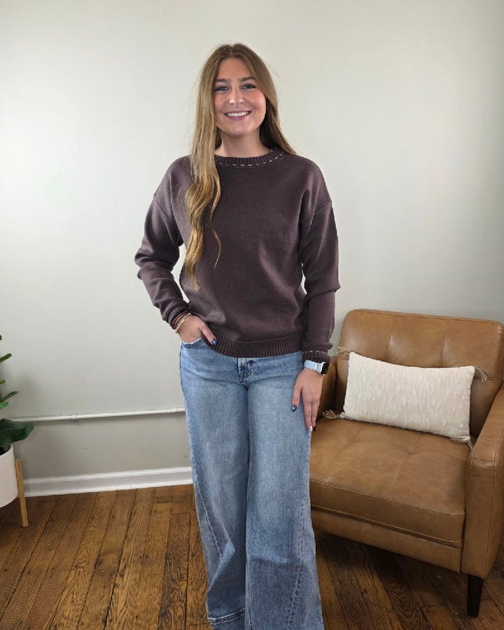 A woman wears a cozy, brown long-sleeved Staccato Willow Stitch Detailed Sweater with decorative neckline stitching, paired with light-wash wide-leg jeans.