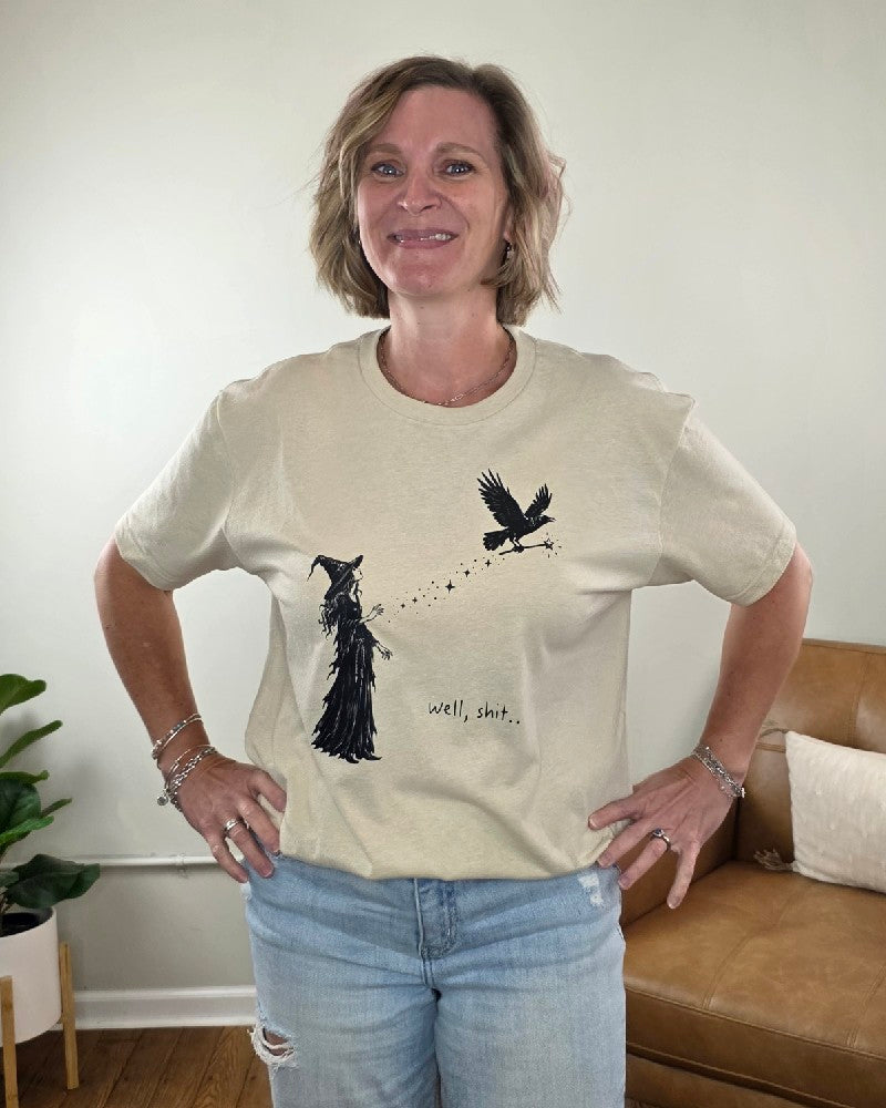 A woman wears a beige Cosmos + Clover Boutique Witch Graphic Tee featuring a witch pointing her wand at a crow, with the text well, shit.. printed below.
